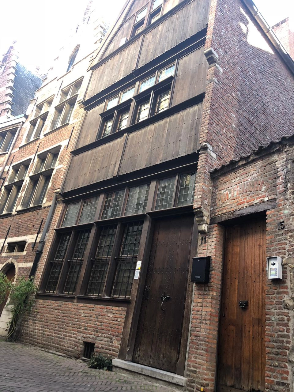 A street-level, low-angle photo of an old European row house. The ground floor is red brick with a large, slanted wooden door and a row of leaded glass windows. The upper facade is covered entirely in vertical dark wooden planks, typical of historic Flemish architecture. A cobblestone street is visible in the foreground.