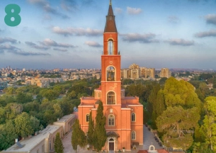 An aerial view of a large, red-brick church with a tall, narrow spire dominating a residential area, likely in a city like Tel Aviv or Jaffa. The church is surrounded by green trees and lower buildings against a sunset or twilight sky. This is likely the Russian Orthodox Church of Tabitha in Tel Aviv-Jaffa.