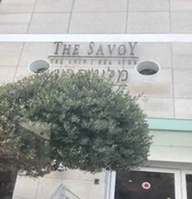 A close-up view of the sign for "THE SAVOY" hotel, engraved in stone on the upper facade of a modern building. Below the English name, the name is also written in Hebrew. The view is partially obstructed by the leaves of a large, manicured shrub.