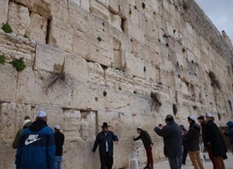 A view of the Western Wall (Kotel) in Jerusalem, showing its large, ancient limestone blocks. Several men, some wearing Jewish religious attire, are gathered at the base of the wall praying or reflecting. The sky is overcast.