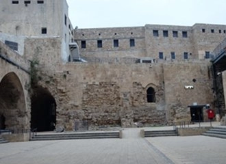 A view of the Crusader fortress walls in the city of Acre (Akko), Israel. The ancient stone walls show multiple layers of construction with small windows and a large arched opening on the left, surrounding a paved, open courtyard.