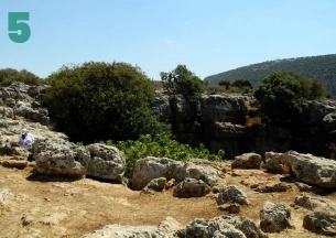 A sunny landscape of a rocky, overgrown plateau with large, scattered limestone boulders and dense green trees. In the distance, a steep, forested hillside rises, with a figure in light robes partially visible in the lower left corner. This location is likely in the Upper Galilee or Golan Heights region.