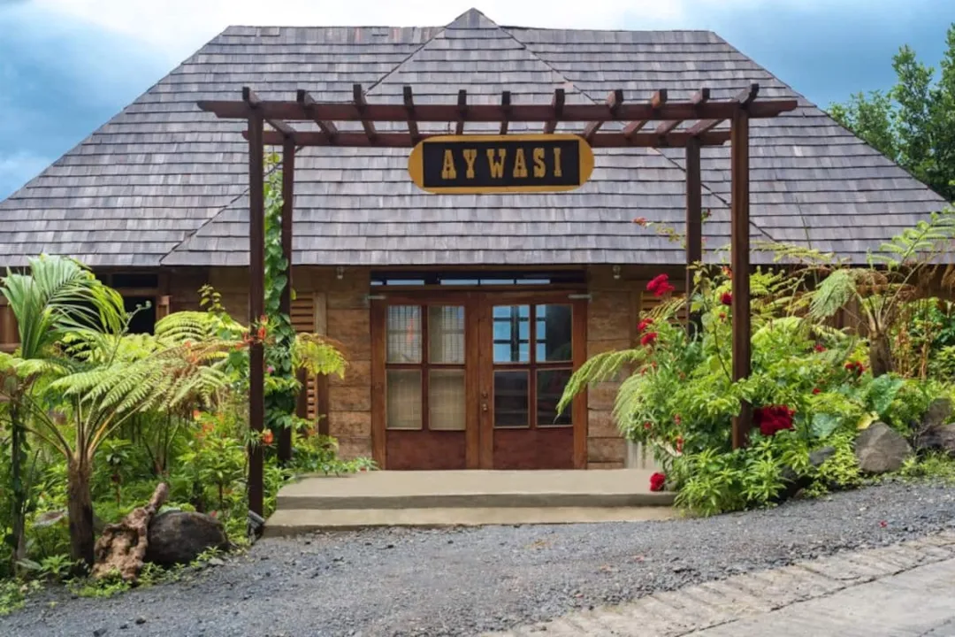 The exterior entrance of a lodge or cottage named "AYWASI." The building has a large, gray wooden shingled roof and stone walls. The front door is framed by a wooden pergola structure with a sign hanging overhead. The entrance is surrounded by lush tropical vegetation, including red flowers and ferns.