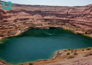 A wide shot of a deep, open-pit mine that has filled with water, creating a vibrant turquoise/green lake surrounded by steep, stepped walls of reddish-brown rock and earth.