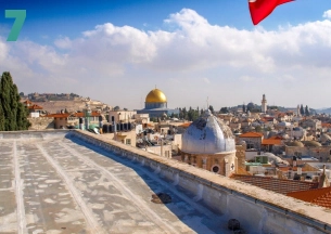 A panoramic view of the Old City of Jerusalem skyline taken from a rooftop. The iconic golden Dome of the Rock is centered in the background, with a silver-domed building in the middle ground, surrounded by densely packed stone buildings with a red flag visible on the right.