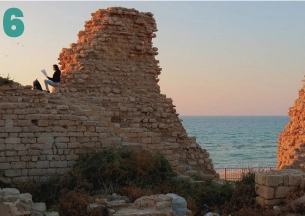 A sunlit view of the massive, ruined stone walls of an ancient coastal fortress. A figure is sitting on the top of the wall, looking out at the sea, which is visible through a gap in the ruins. The light suggests sunrise or sunset. This is likely the Roman ruins at Caesarea Maritima.