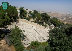 An aerial view of a large, stone outdoor amphitheater, or tiered seating area, set into a hillside surrounded by lush green pine trees and foliage. Buildings are visible on the hill above, and a hazy valley view is in the background.