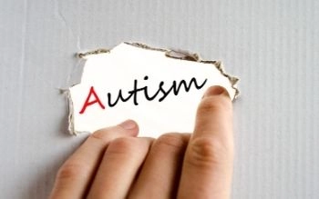 A close-up shot of a hand peeling back a torn piece of light gray material to reveal the word "Autism" written in black script underneath on a white surface. The letter 'A' in Autism is stylized in red.