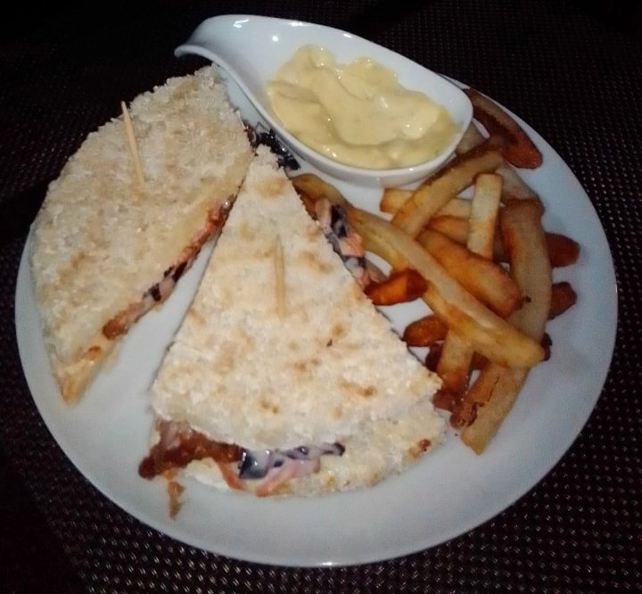 A white plate holds a meal consisting of two triangular sandwich halves with a crispy, breaded exterior and filling, served with a side of golden-brown french fries and a small, white, boat-shaped dish of creamy dipping sauce or mayonnaise.