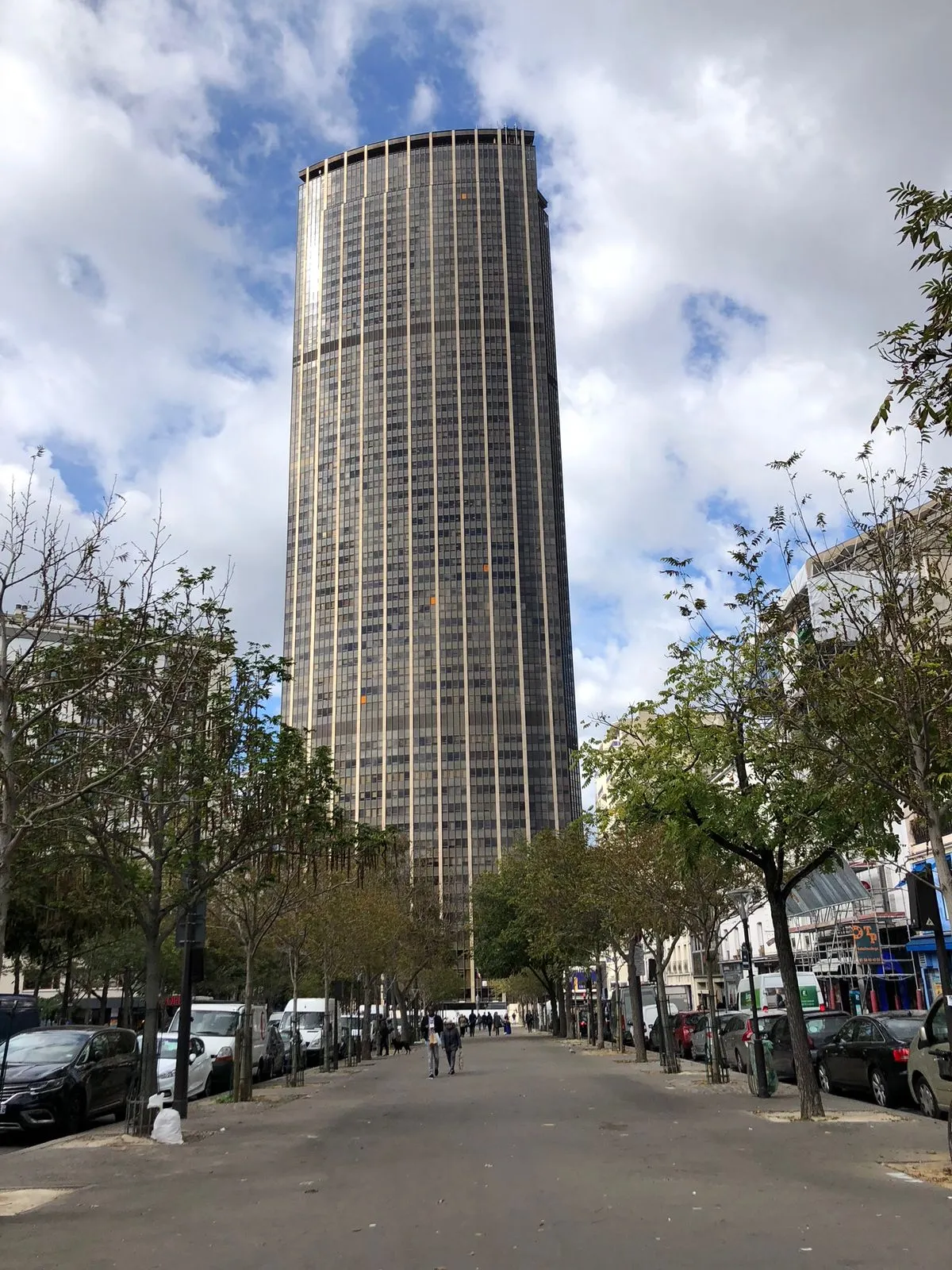 A street-level vertical photo of the Montparnasse Tower (Tour Montparnasse), a tall, dark-glass skyscraper in Paris. The building dominates the background, rising straight up under a cloudy blue sky. In the foreground, there is a wide pedestrian walkway or street lined with small trees and parked cars.