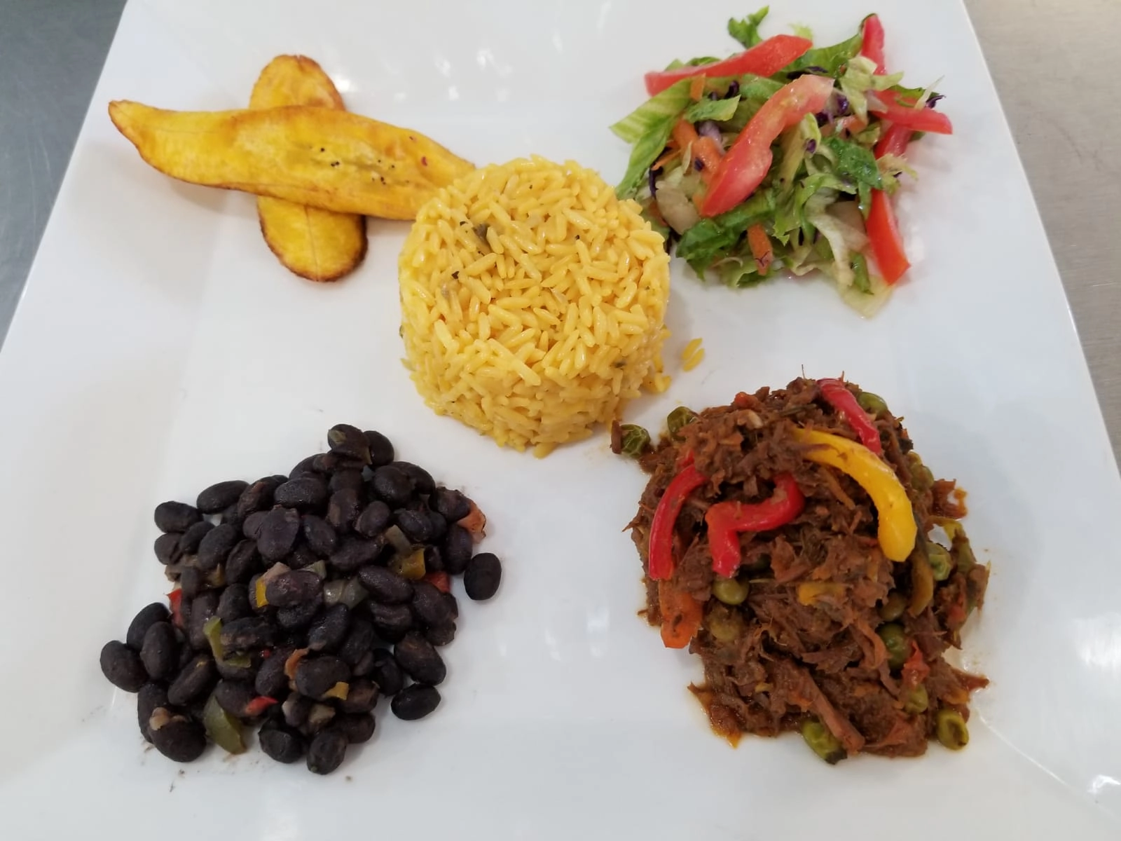 A square white plate holding a meal with four distinct components: a mound of yellow rice, a pile of black beans seasoned with vegetables, a portion of shredded stewed meat (possibly Ropa Vieja) mixed with peas and colorful bell peppers, and two pieces of fried plantain (tostones or maduros) with a small side salad.
