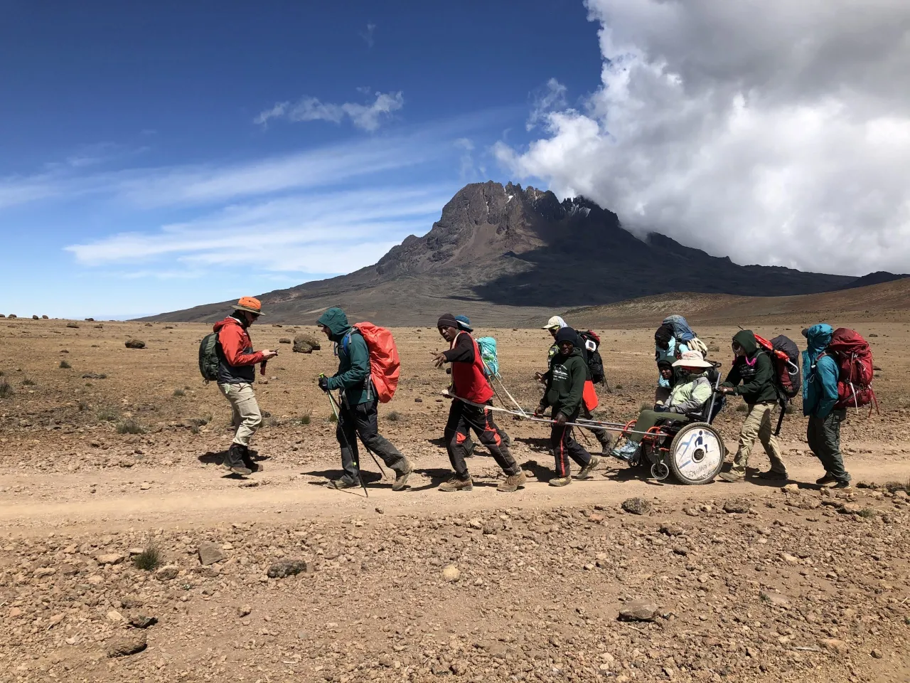 A group of seven hikers wearing heavy backpacks and hiking gear traverse a wide, dusty, brown, flat plain under a bright blue, cloudy sky. They are collaboratively pushing and pulling a specialized, large-wheeled outdoor wheelchair carrying a passenger. In the background, a large, dramatic, rocky mountain peak, partially obscured by clouds, dominates the horizon.