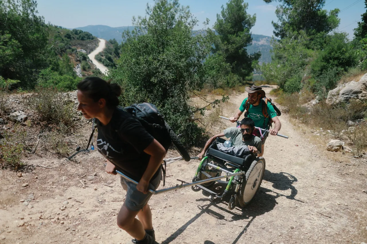 A man in a specialized, rugged, single-wheel outdoor chair is pushed and pulled by two hikers on a wide, sunny dirt trail. A woman with dark hair in a black shirt pulls from the front using metal handles, and a man in a green shirt and sun hat pushes from the back. A winding road is visible in the distance on the hillside, surrounded by green trees.