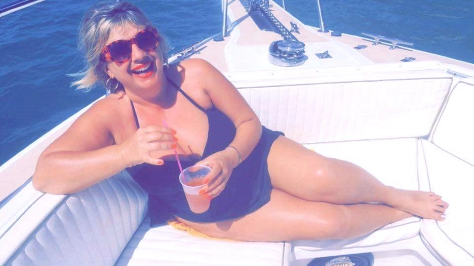 A smiling woman with blonde hair and red sunglasses lounges on the white cushioned seating of a boat deck on a sunny day. She is wearing a dark swimsuit and holding a pink-orange drink with a straw. The ocean water is visible in the background.