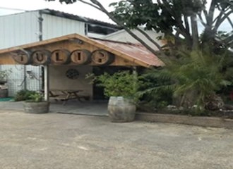 The exterior entrance to a winery or restaurant called "TULIP." The sign is made of wooden barrels with individual letters painted on them. The entrance is covered by a wooden roof and surrounded by trees and large planters, with a picnic table visible underneath.