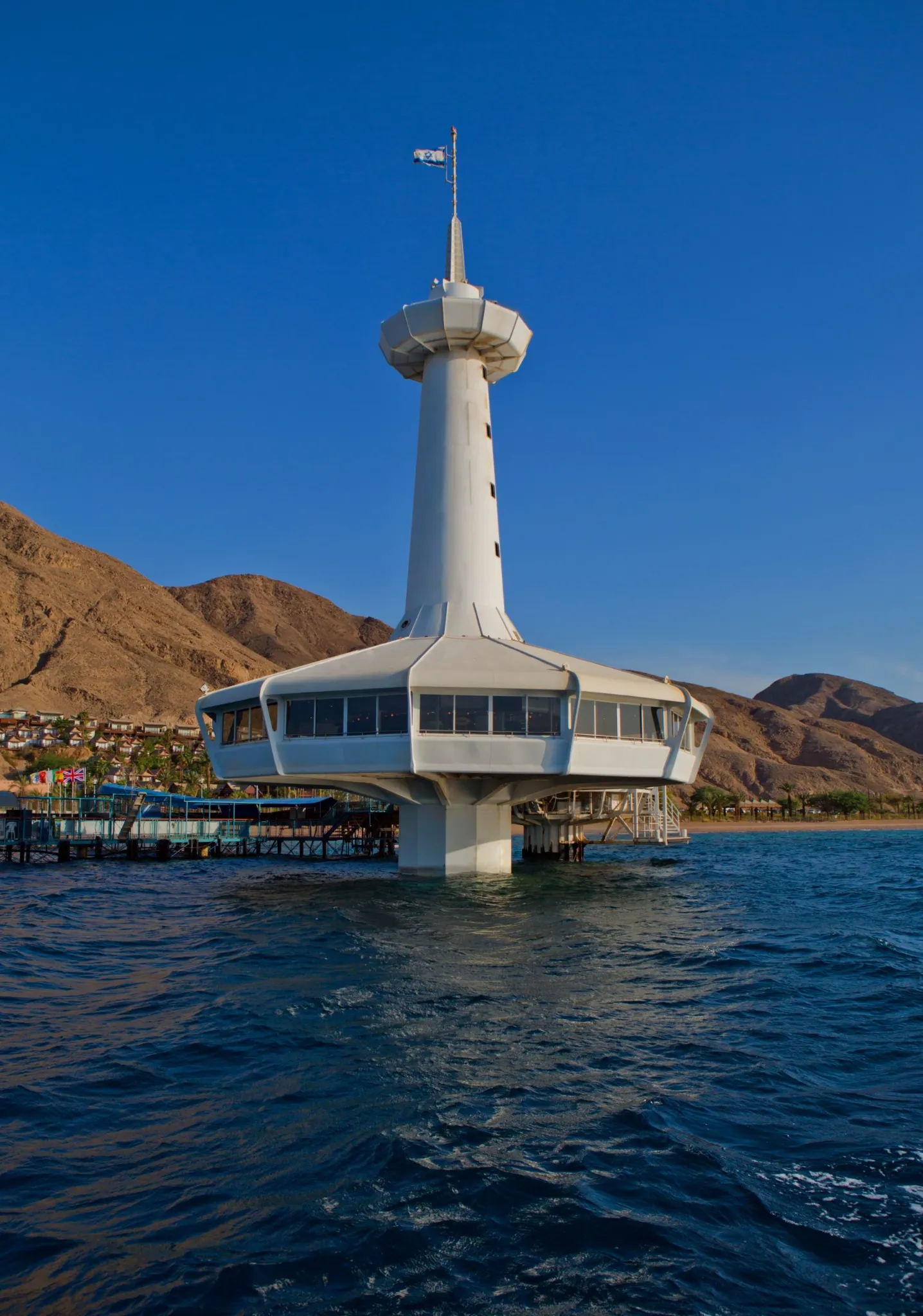A prominent, futuristic-looking, white tower structure, likely the Coral World Underwater Observatory, stands in the water near the shore. The structure features a wide observation deck and a tall central tower, backed by steep, reddish-brown mountains and a bright blue sky.