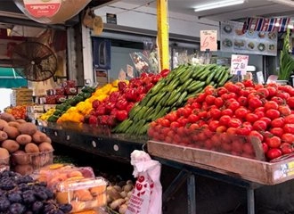 A vibrant display of fresh produce at an indoor market stall. The display features rows of ripe red tomatoes, green cucumbers, red bell peppers, and yellow bell peppers. Baskets of kiwis and plums are visible in the foreground.