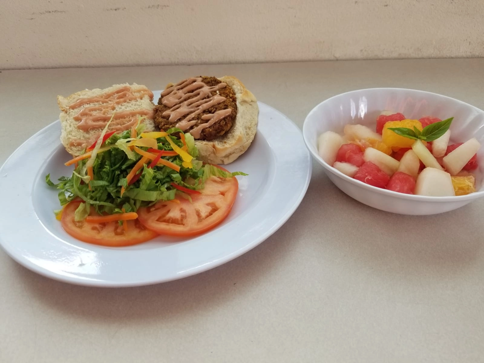 A white plate with an open-faced sandwich consisting of a toasted bun and a veggie or meat patty drizzled with a light sauce. Beside the sandwich is a small salad of lettuce, shredded carrots, and sliced tomatoes. To the right is a white bowl of diced fresh fruit salad, including watermelon, melon, and citrus segments, topped with a mint or basil lea