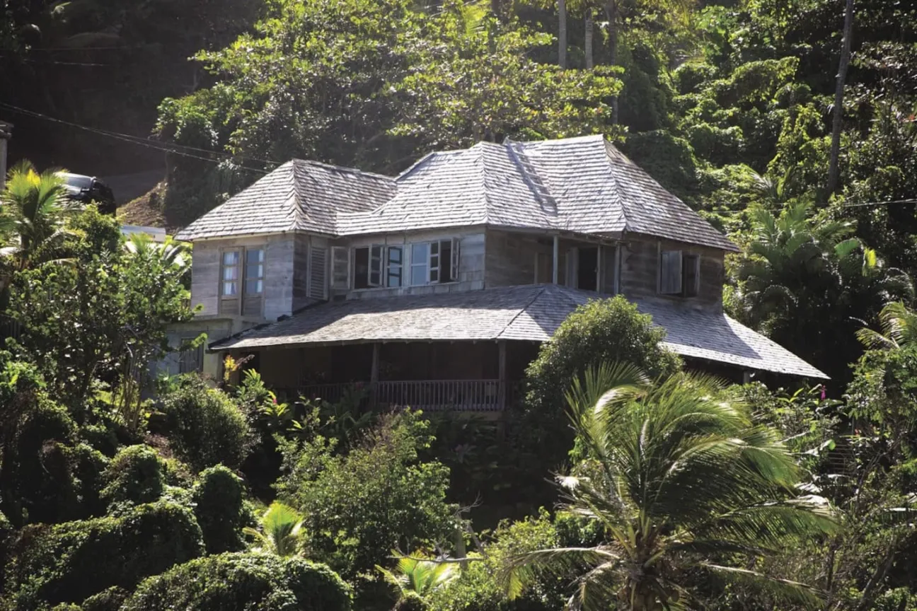 A two-story, traditional-style wooden house with a light gray shingle roof and a wrap-around porch is nestled high on a lush, steep hillside, almost completely surrounded by dense, vibrant green tropical foliage and trees. The house has several windows with open shutters, suggesting a sunny day.