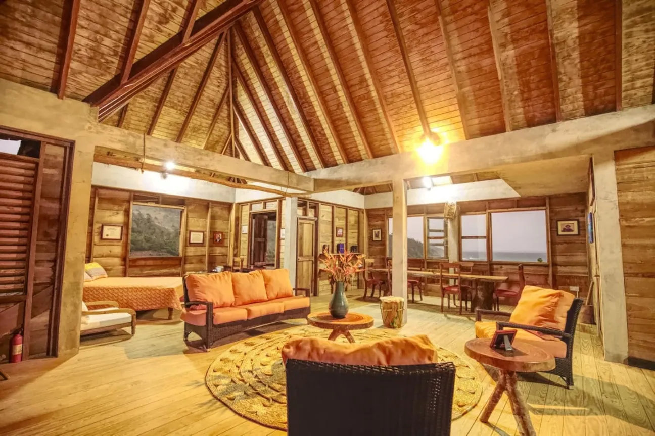 The interior of a spacious, open-plan living area with a high, vaulted wooden ceiling and wood-paneled walls and floors. The room is furnished with orange-cushioned wicker sofas and chairs arranged around a round, textured rug. The space also includes a dining area and a visible bed in the far left, with windows offering views of the exterior landscape.