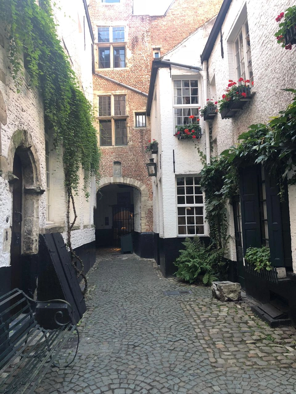 A narrow, charming cobblestone alleyway in a historic European city. The buildings on either side are painted white with black trim and are adorned with climbing ivy, window boxes of red flowers, and dark shutters. The path leads toward a red brick building and a dark, arched passageway in the distance. A dark green metal bench is visible in the lower left corner.