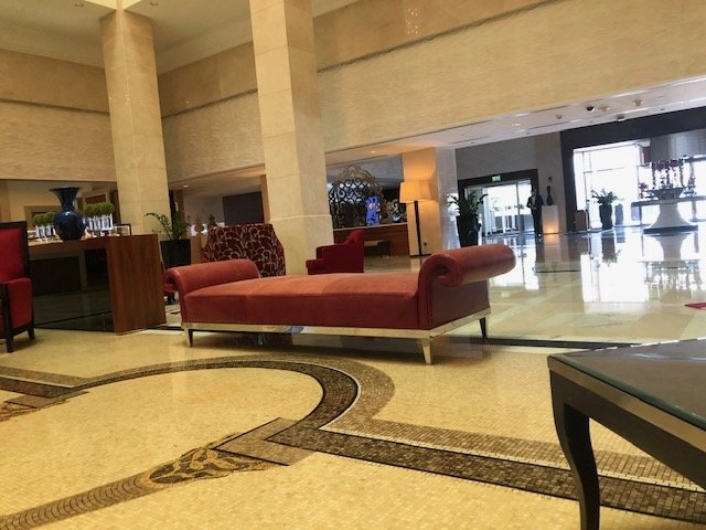 The luxurious, brightly-lit lobby of a hotel featuring a large, red, velvet chaise lounge in the center of the tiled floor. The floor has an ornate, dark mosaic pattern. Tall, cream-colored pillars and glass doors leading outside are visible in the expansive space.