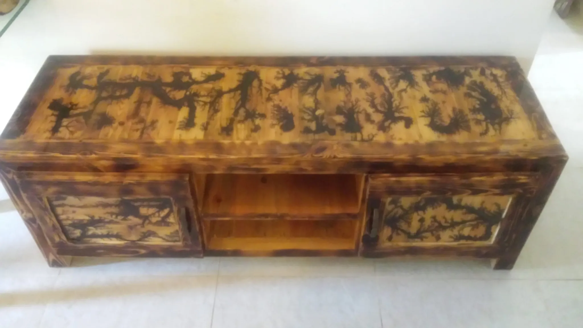 A rectangular wooden cabinet, possibly an entertainment unit or dresser, with a natural wood stain. The top surface and the two cabinet doors feature dark, intricate, branching burn patterns created using the Lichtenberg figure technique. The center has two open shelves.