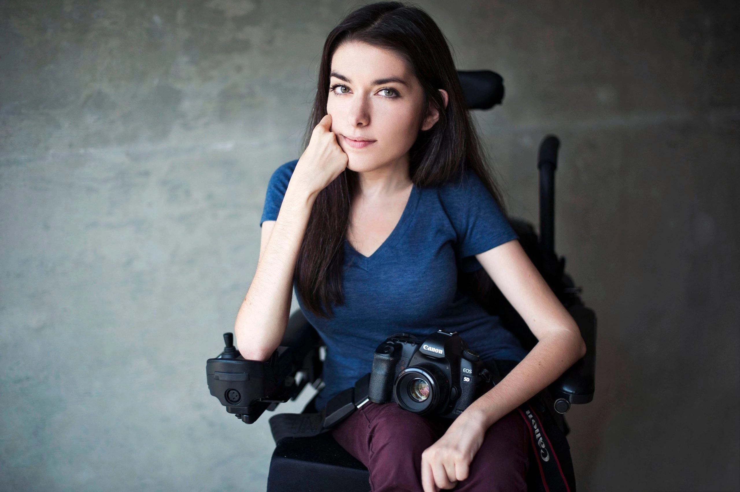A young woman with long dark hair, wearing a blue V-neck shirt and burgundy pants, is seated in a power wheelchair and looks directly at the camera. She is resting her chin on her hand, and an SLR camera is resting on her lap. The background is a plain gray concrete wall.
