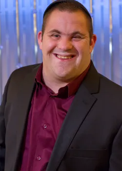 A portrait of a young man with a dark skullcap (kippah) wearing a dark blazer over a burgundy dress shirt, smiling broadly at the camera. He is standing in front of a shimmering blue and white vertically striped background.