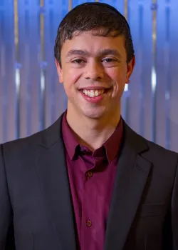 A portrait of a man with Down syndrome wearing a dark blazer over a burgundy dress shirt, smiling brightly at the camera. He is wearing a dark skullcap (kippah) and is standing in front of a shimmering blue and white vertically striped background.