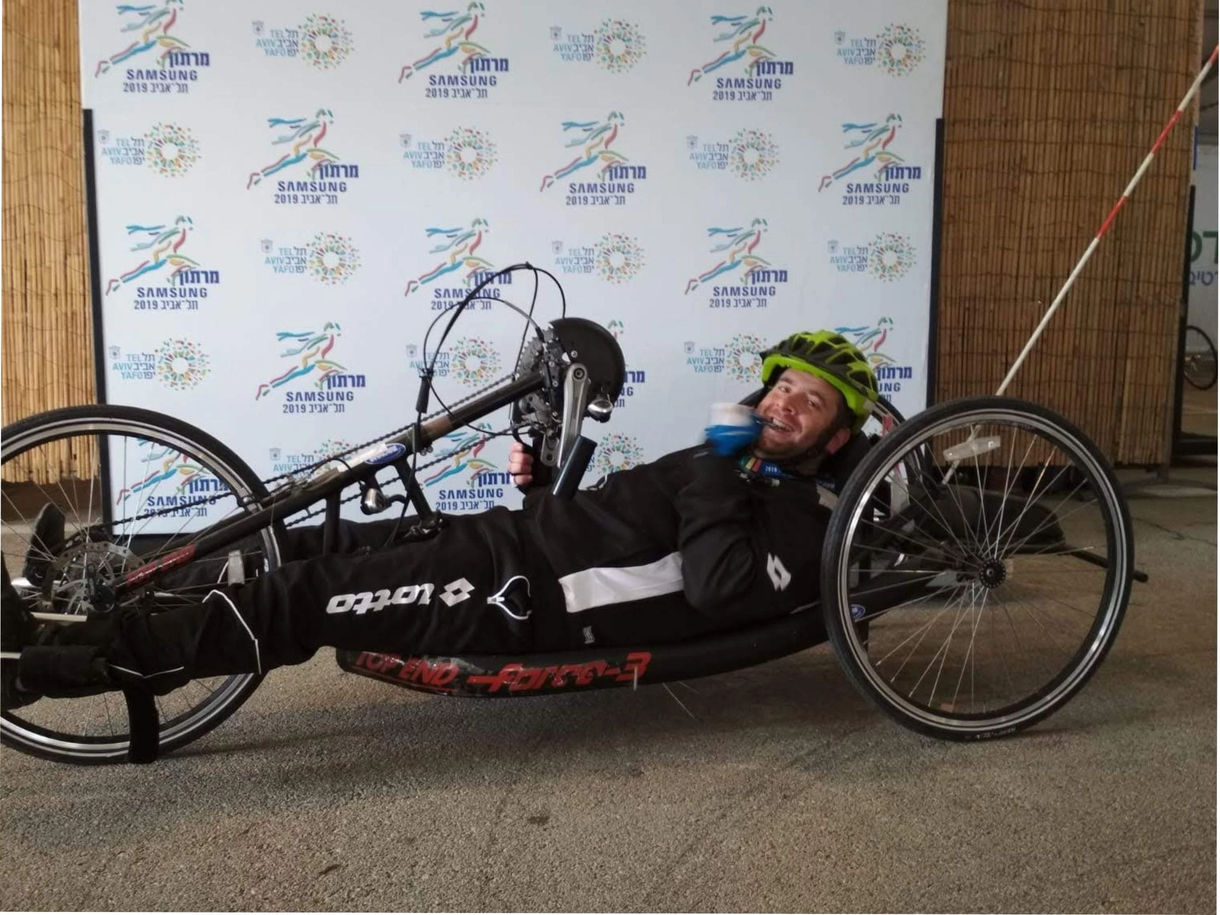 A man lying in a three-wheeled racing handcycle (hand bike), smiling and holding a water bottle. He is wearing a black racing suit with a Lotto logo and a bright green helmet. The backdrop is a repeating banner for the "Samsung Tel Aviv 2019 Marathon," featuring the event logo and text in Hebrew.