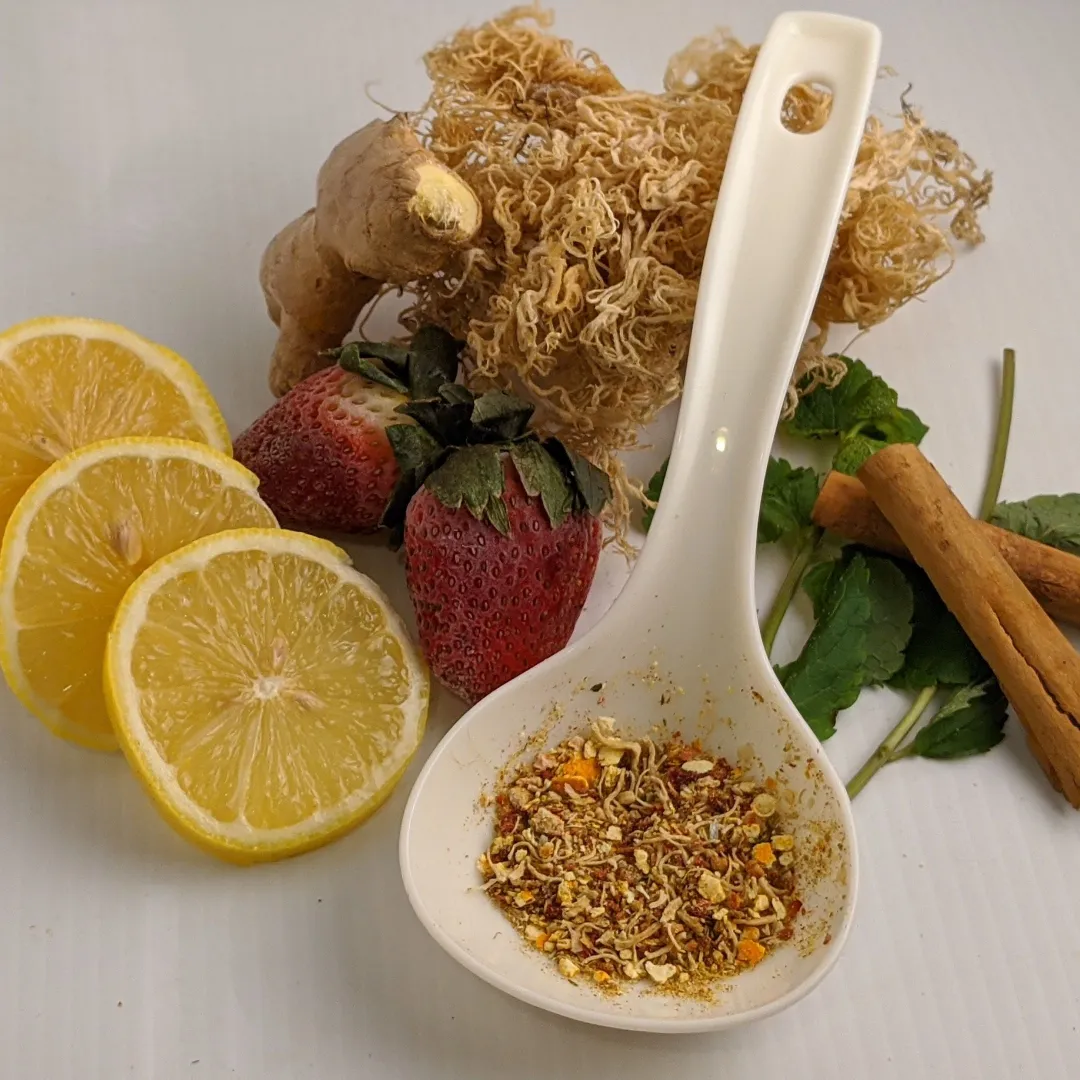 A close-up still life photograph of various health and food ingredients arranged on a white surface. The items include three slices of lemon, two strawberries, a piece of ginger root, a clump of dried sea moss, two cinnamon sticks, and a sprig of mint. In the foreground, a white ceramic spoon holds a small mixture of dried spices or seasonings, including chili flakes and yellow specks.