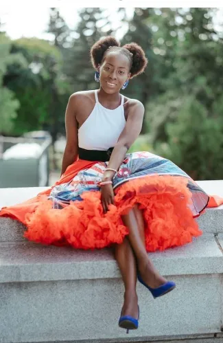 A young Black woman sits outdoors on a stone ledge, smiling directly at the camera. She is wearing a white halter top and a vibrant, full skirt with a striking pattern and voluminous orange tulle underskirt. She has blue earrings and blue high-heeled shoes. Her hair is styled in two afro puffs.