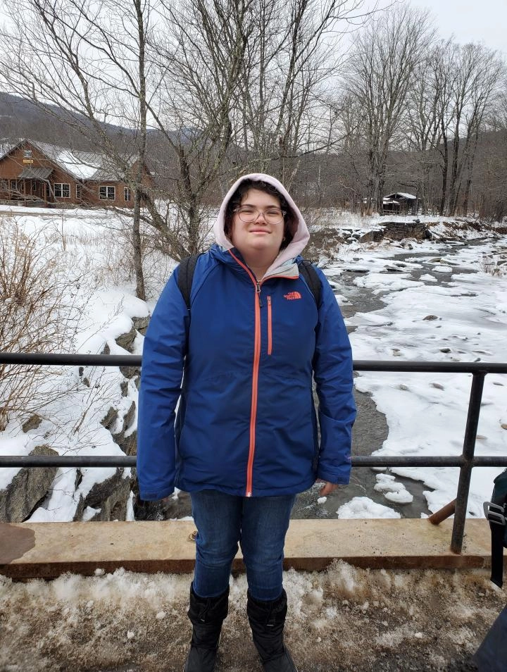 A person wearing a blue North Face jacket with an orange zipper, a pink hooded sweatshirt, and jeans, stands on a bridge over a partially frozen river in a snowy landscape. The person is looking straight ahead, smiling slightly and wearing glasses. In the background, a wooden cabin building is visible through leafless trees.