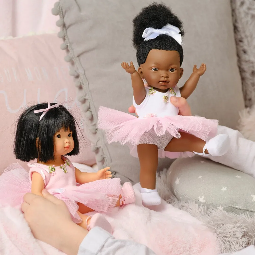 A close-up shot of two baby dolls dressed as ballerinas, positioned on a soft, pink, and grey-pillowed surface. On the right, a doll with dark skin and a high Afro puff tied with a white bow is held up in a ballet pose, wearing a white leotard, a pink tulle tutu, and white socks. On the left, a doll with lighter skin, dark straight hair tied with a pink bow, is sitting and wearing a light pink leotard, a pink tulle tutu, and pink ballet shoes. Two sets of hands are visible holding the dolls.