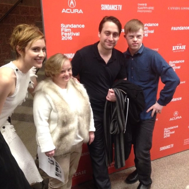 Four people stand together and smile in front of a bright red step-and-repeat backdrop for the Sundance Film Festival 2016. From left to right: a woman in a white dress, a short woman wearing a white fur vest, a man in a black shirt, and a young man with Down syndrome in a blue patterned shirt.