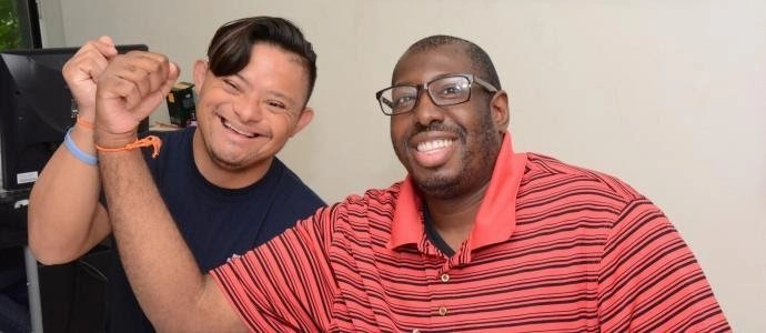 Two men smiling and holding hands in a celebratory gesture. The man on the left has Down syndrome and is wearing a dark shirt. The man on the right is wearing a red and black striped polo shirt and glasses. They are sitting indoors.