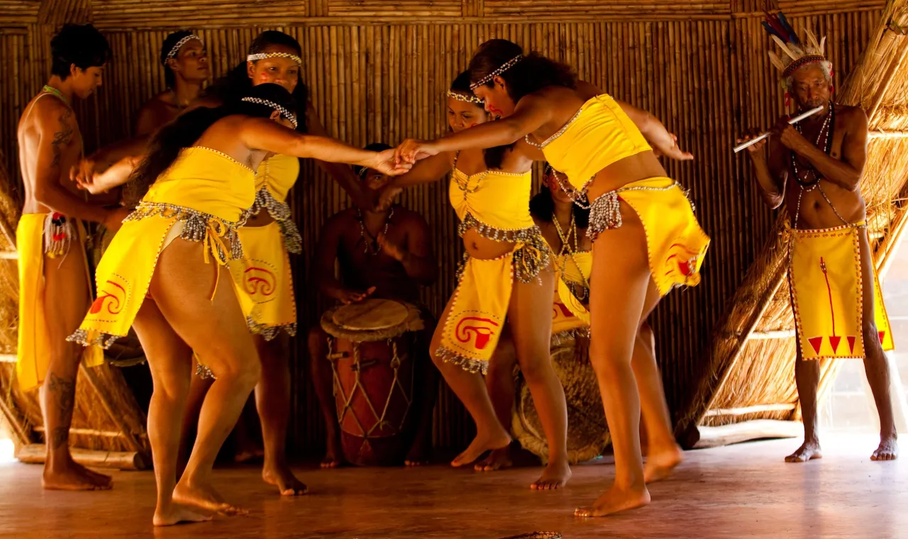 A group of indigenous people performs a traditional dance inside a structure with bamboo walls. Several women, wearing short, bright yellow outfits with beaded trim, are holding hands in a circle. Barefoot men, also in yellow attire, are positioned around them, with one man playing a flute on the right and another sitting and playing a drum in the center background.
