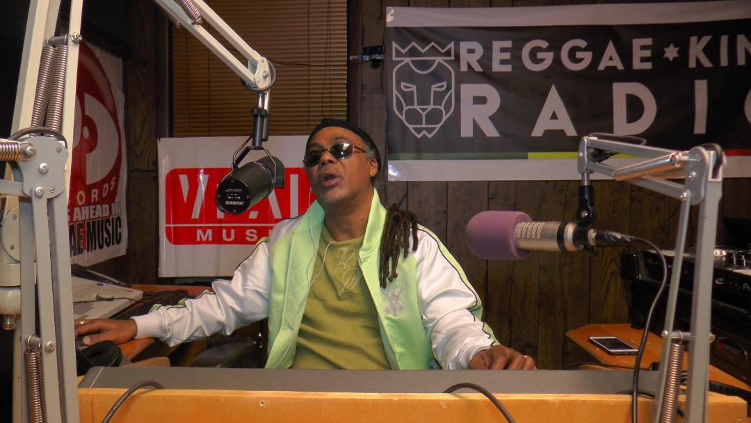 A middle-aged man with dreadlocks, wearing sunglasses, a black beanie, a light green jacket with white trim, and a green shirt, is seated at a radio console in a broadcast booth. He is speaking into a microphone on a boom arm. Promotional signs, including a "REGGAE KING RADIO" banner, are visible on the wall behind him.