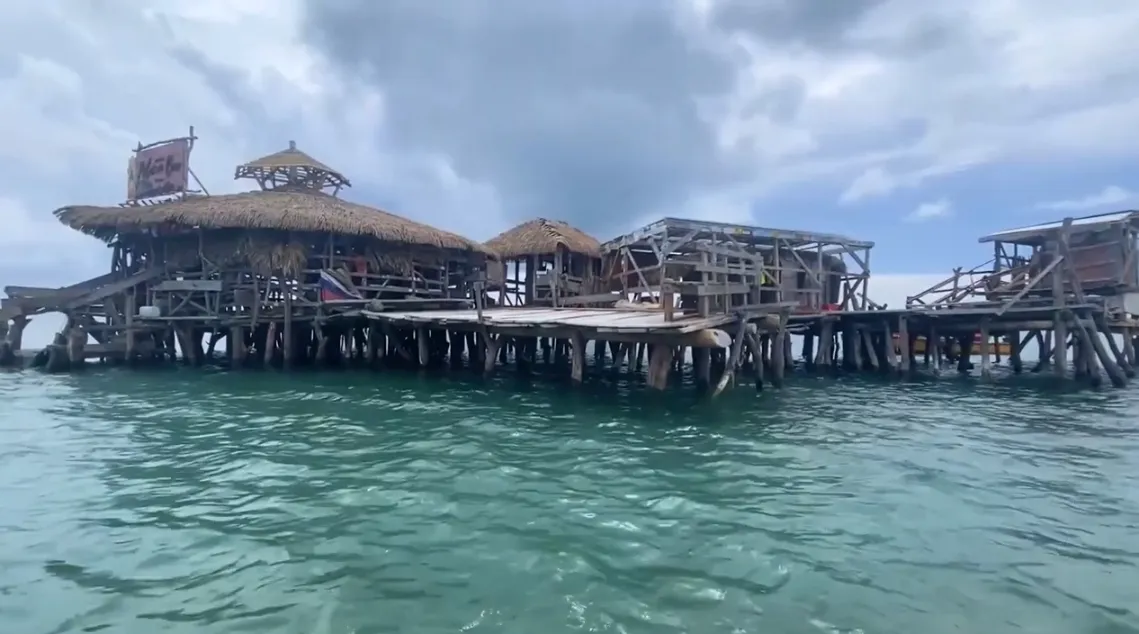 A wide shot of a rustic wooden structure built on stilts over the ocean water, possibly a bar or restaurant. The structure features thatched roofs and exposed wooden beams and decking, standing under a cloudy sky.