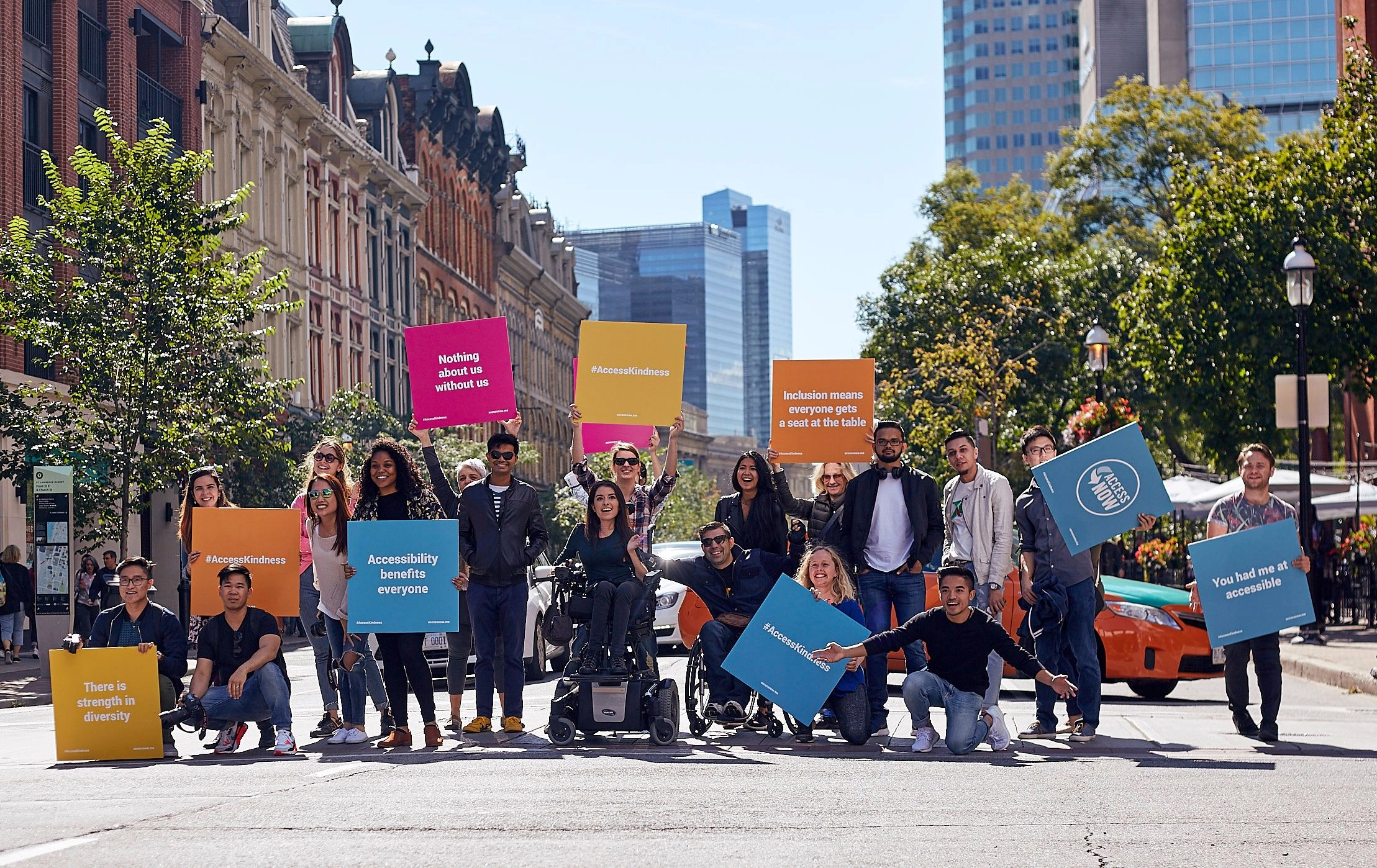 A diverse group of people, including two individuals in wheelchairs, stand in a sunny city street holding colorful square signs with accessibility and inclusion slogans. Slogans include 'Accessibility benefits everyone' and 'Nothing about us without us.'