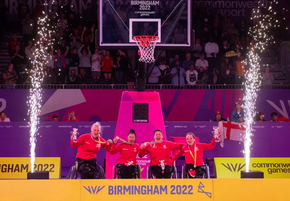 A celebratory photo of a four-person Wheelchair 3x3 Basketball team from Wales on a podium. The four athletes, all women in red jerseys and seated in their wheelchairs, are holding up medals and teddy bears while fireworks or pyrotechnic fountains erupt on either side of them. The background shows spectators and a scoreboard that reads "BIRMINGHAM 2022 Commonwealth Games."