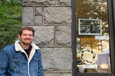 A head-and-shoulders portrait of a smiling young man with a beard and curly brown hair. He is wearing a dark denim jacket with a white sherpa collar over a plaid shirt. He is standing outdoors, leaning against a rough-cut stone wall with green trees visible on the left.