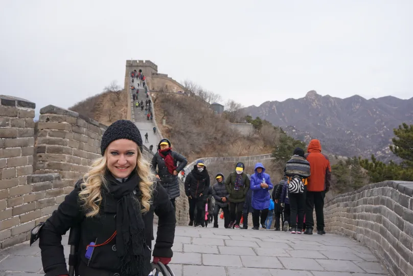 Person smiling in front of a section of the Great Wall of China, wearing a black beanie and scarf. The wall stretches up a hill with several other visitors climbing the path, bundled up in winter clothing. The surrounding landscape has barren trees and mountainous terrain under a gray sky. (China, Great Wall, November 2017)