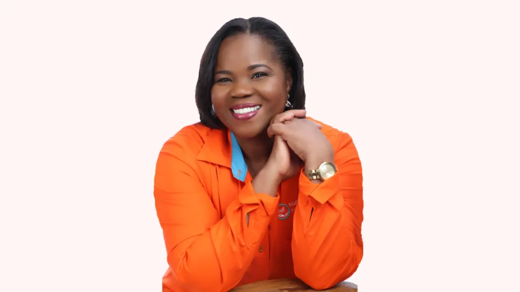 A professional portrait of a smiling Black woman wearing a bright orange button-down shirt with a light blue collar. She is resting her chin on her clasped hands and is wearing a gold wristwatch. The background is a plain white/light pink gradient.