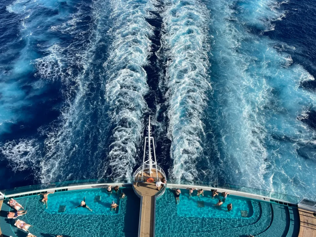 A stunning, high-angle view from the back of a cruise ship showing the powerful white wake tracks cutting through the deep blue ocean water. Below, the ship's stern features a unique infinity pool with passengers swimming and lounging, overlooking the sea.
