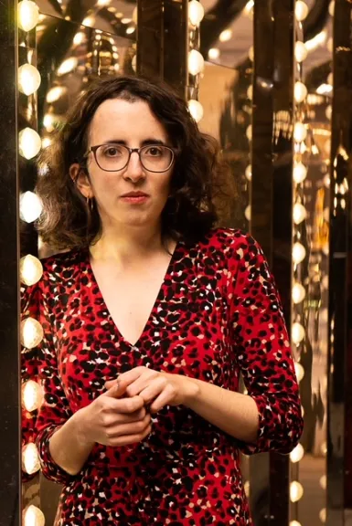 A portrait of a woman, possibly author Lydia Wilkins, wearing glasses and a red and black leopard-print dress with a V-neck. She is standing indoors in front of a reflective surface illuminated by vertical strips of warm, glowing lights.