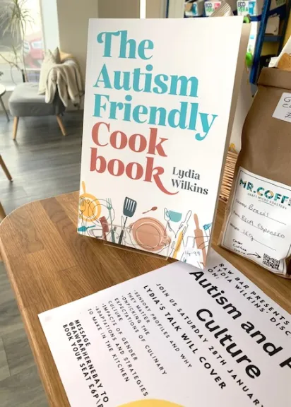 A copy of the book "The Autism-Friendly Cookbook" by Lydia Wilkins standing upright on a wooden table, next to a flyer advertising "Lydia Wilkins' talk" on "Autism and Culture." A bag of coffee is also visible to the right, and the background is a blurred cafe or bookstore setting.