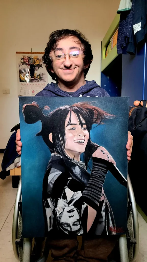 A young man with dark curly hair and glasses, seated in a wheelchair, holding up a large, vibrant oil painting of Billie Eilish smiling and holding a microphone against a dark blue background.