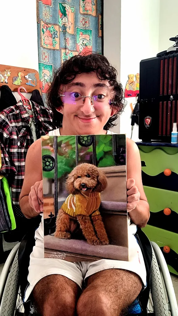 A young man with dark curly hair and glasses, seated in a wheelchair, holding up a realistic painting of a fluffy brown poodle wearing a yellow sweater, sitting on stone steps outdoors.